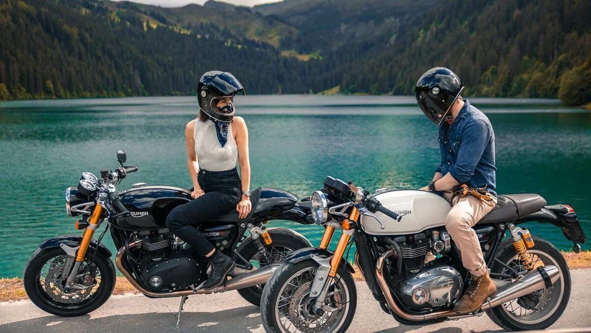 Uma mulher em uma moto preta e um homem em uma moto branca perto de um lago