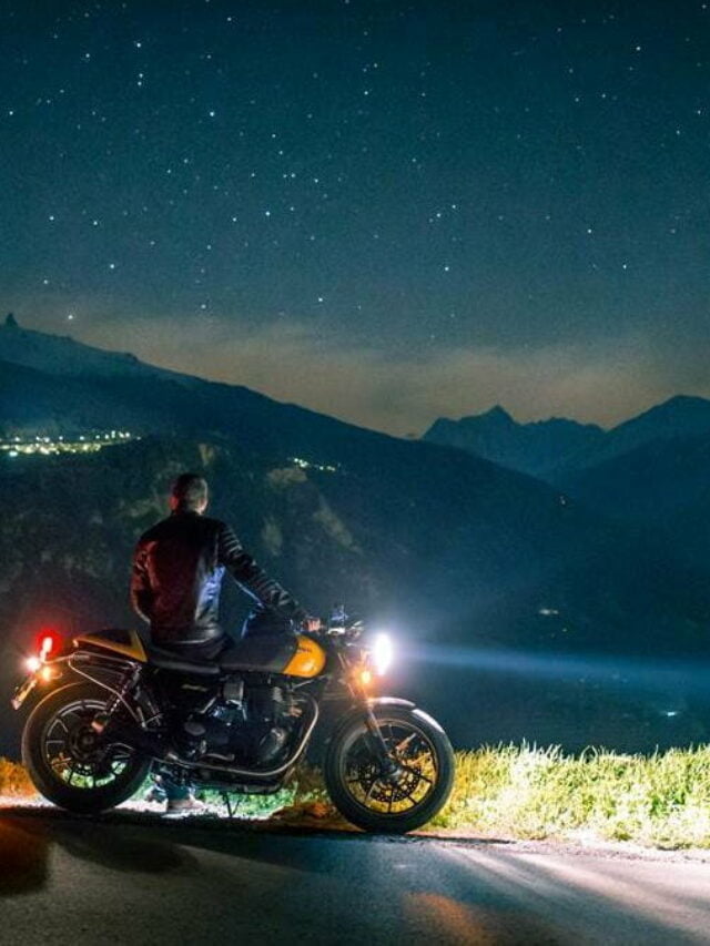 motoqueiro olhando para o horizonte, encostado em moto com farol acesso, em uma estrada, à noite