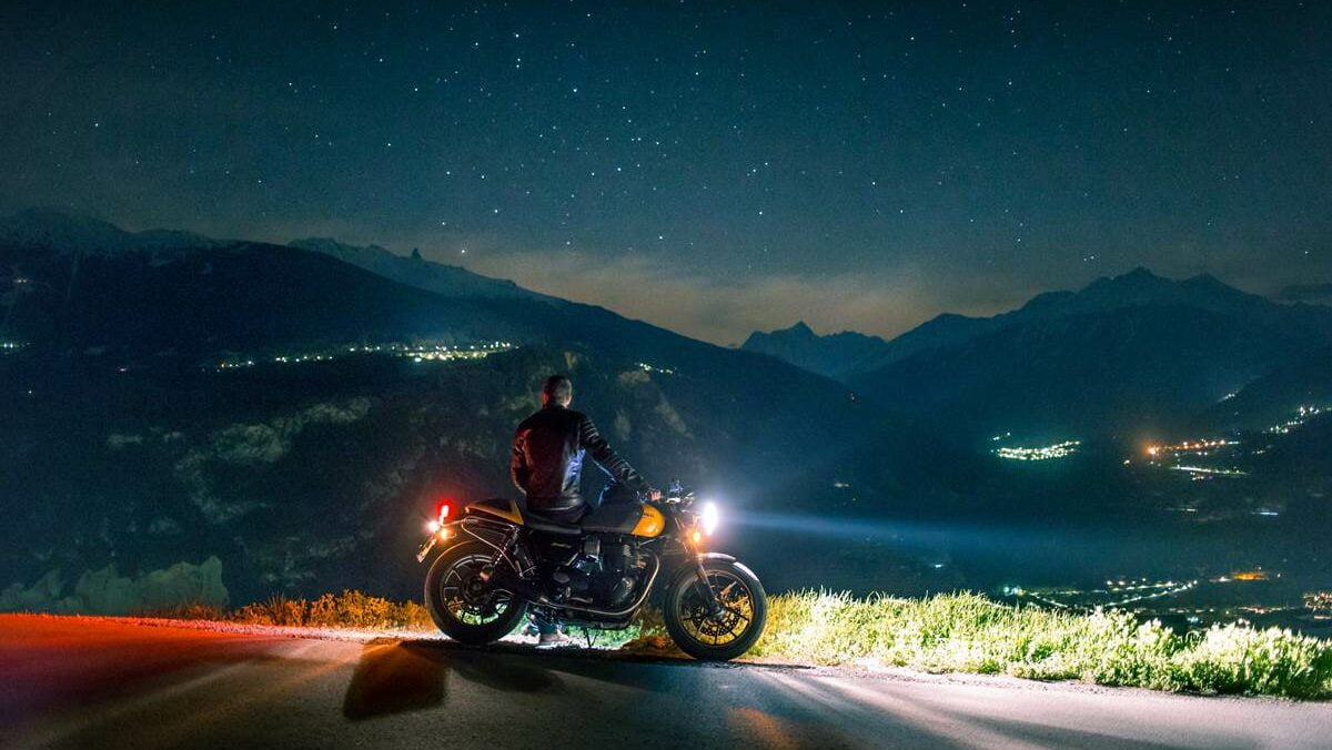 motoqueiro olhando para o horizonte, encostado em moto com farol acesso, em uma estrada, à noite