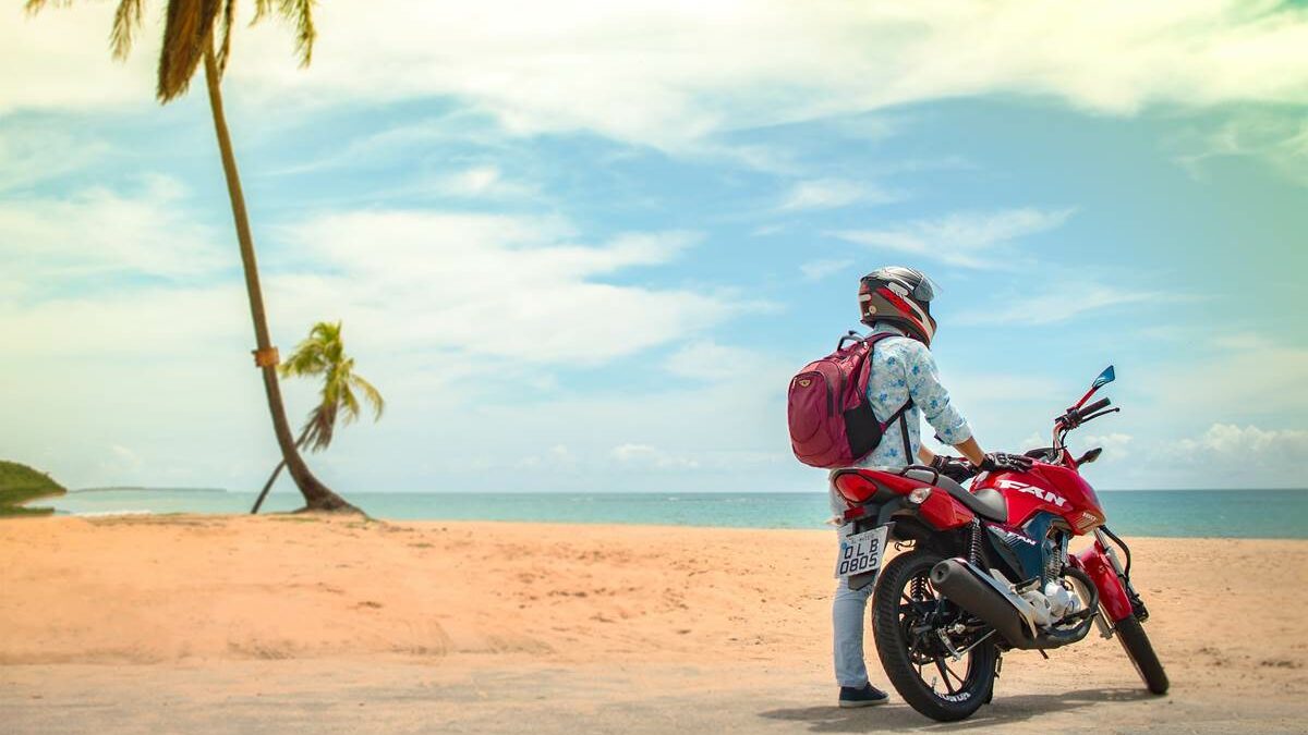 motoqueiro com camisa azul, mochila vermelha, apoiado em moto vermelha em uma praia, em um dia de Sol