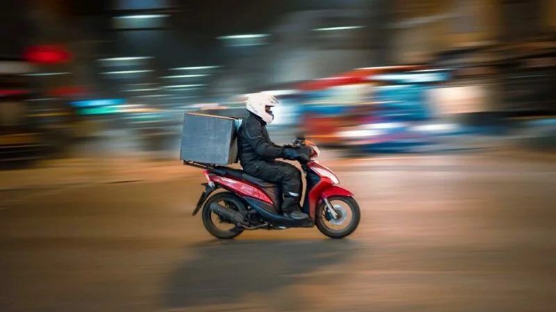 Entregador com roupa preta e capacete branco pilotando uma scooter vermelha