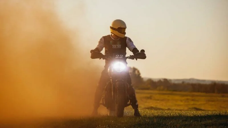 Motoqueiro com capacete branco, em cima de moto, com farol acesso e nevoa de poeira ao lado direito