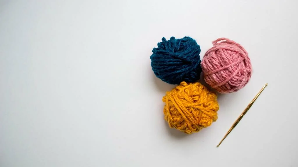 3 novelos de lá, um azul, outro amarelo e um rosa, ao lado de uma agulha